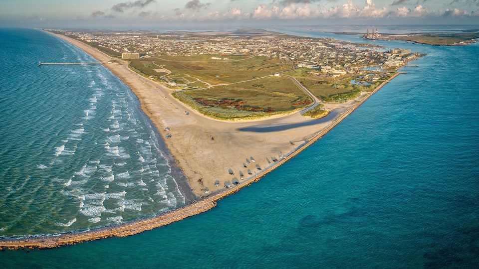 An aerial view of a coastal city with a beach, a breakwater, and a distant industrial structure.