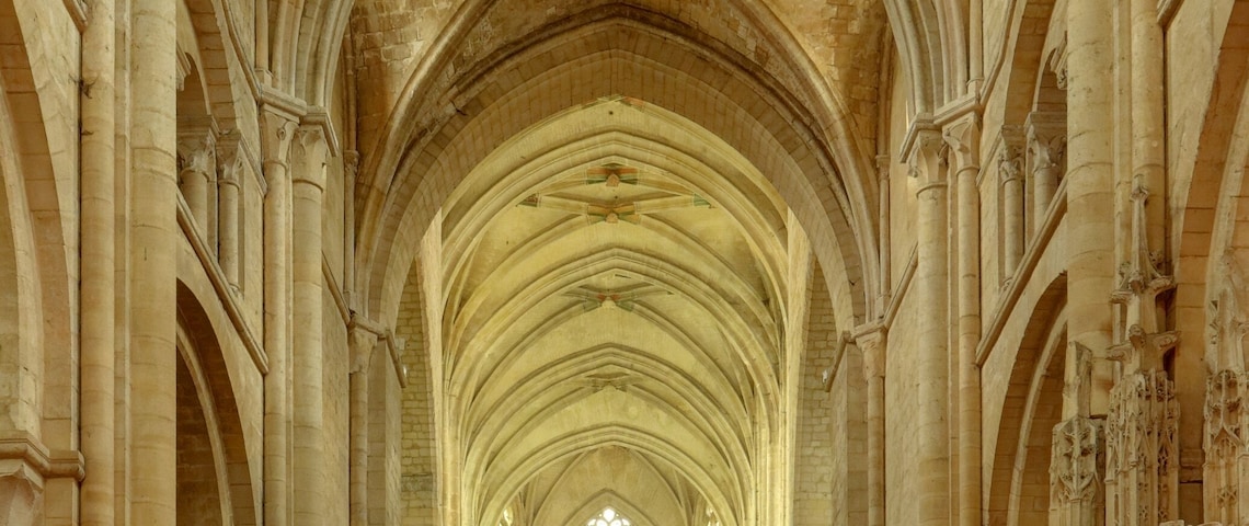Interior de catedral com altos tetos abobadados, fileiras de bancos de madeira e uma nave central que leva a um altar.