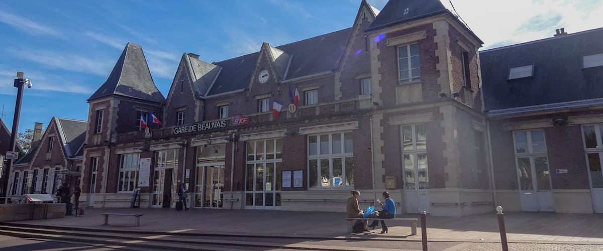 A historic building with a clock tower and flags, a sign reading "GARE DE BEAUVAIRES," and people sitting on benches.
