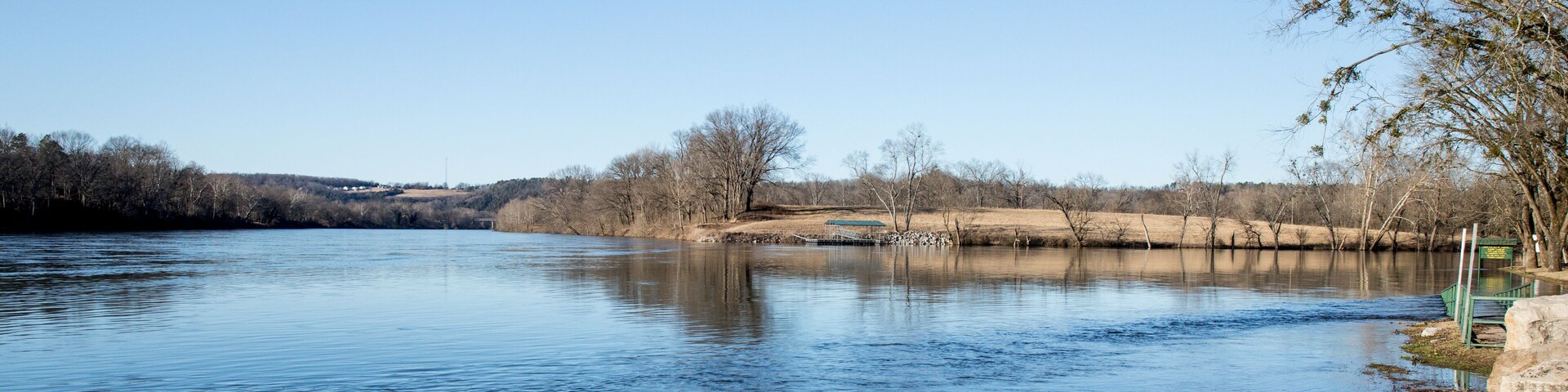 A calm river with clear water reflecting the sky. There are trees and a grassy bank on the right.