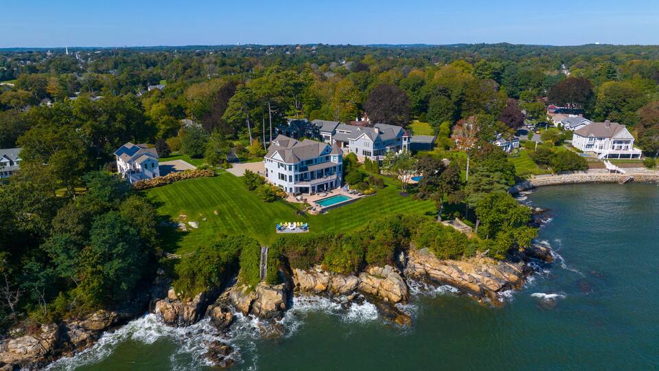 A large estate with a swimming pool, lush green lawns, and a rocky shoreline by the water.