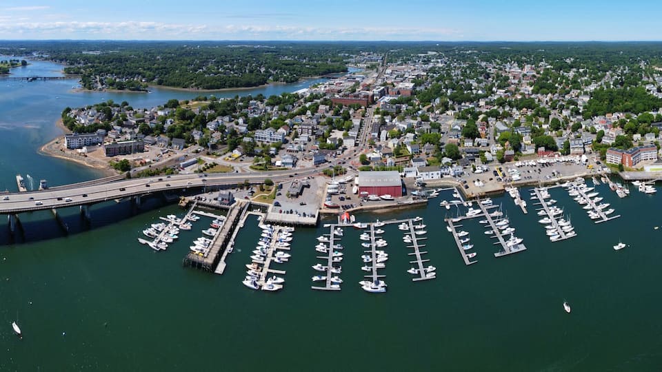 An aerial view of a coastal town with a marina, a bridge, and surrounding greenery.
