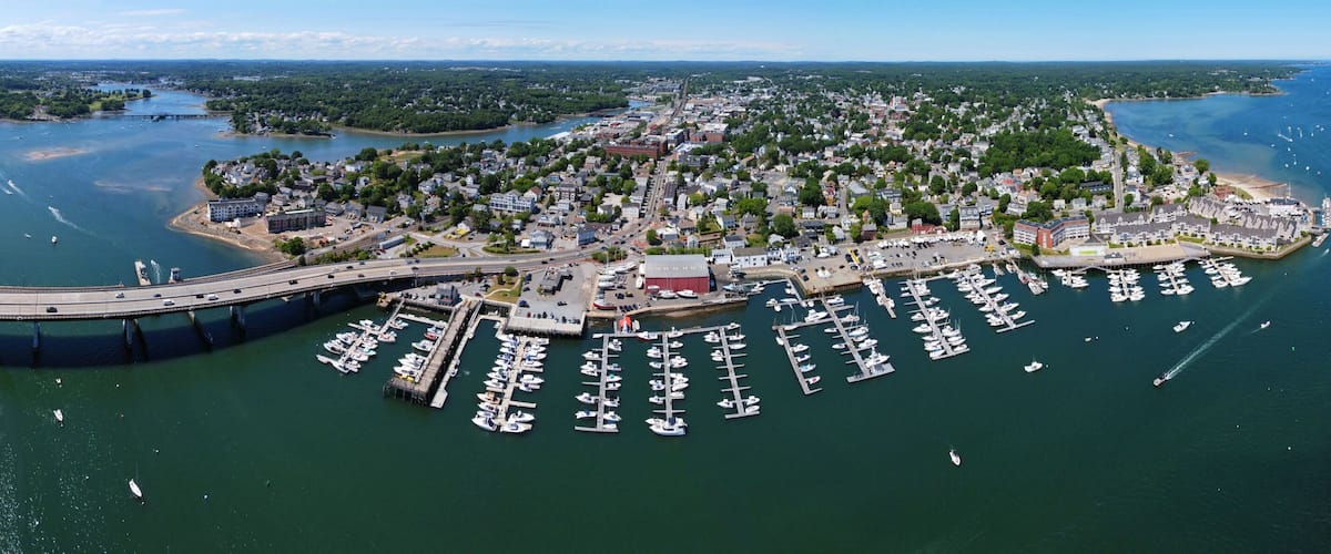 An aerial view of a coastal town with a marina, a bridge, and surrounding greenery.