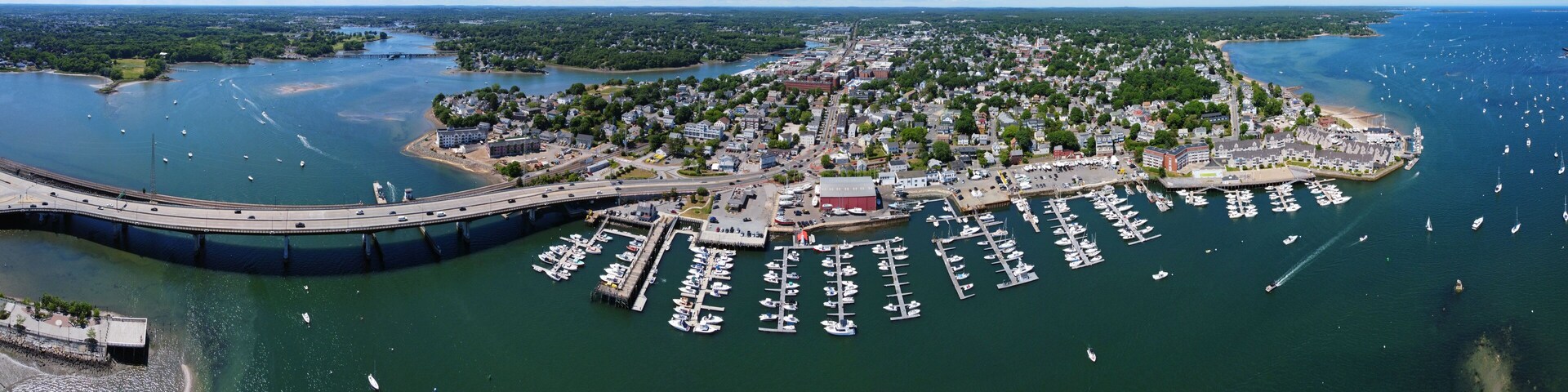 An aerial view of a coastal town with a marina, a bridge, and surrounding greenery.