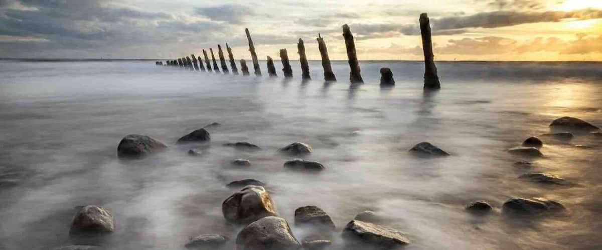 A beach with a row of vertical wooden posts and scattered rocks under a cloudy sky.