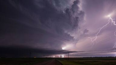 A rural landscape with a dirt road leading to a distant city, under a stormy sky with lightning.