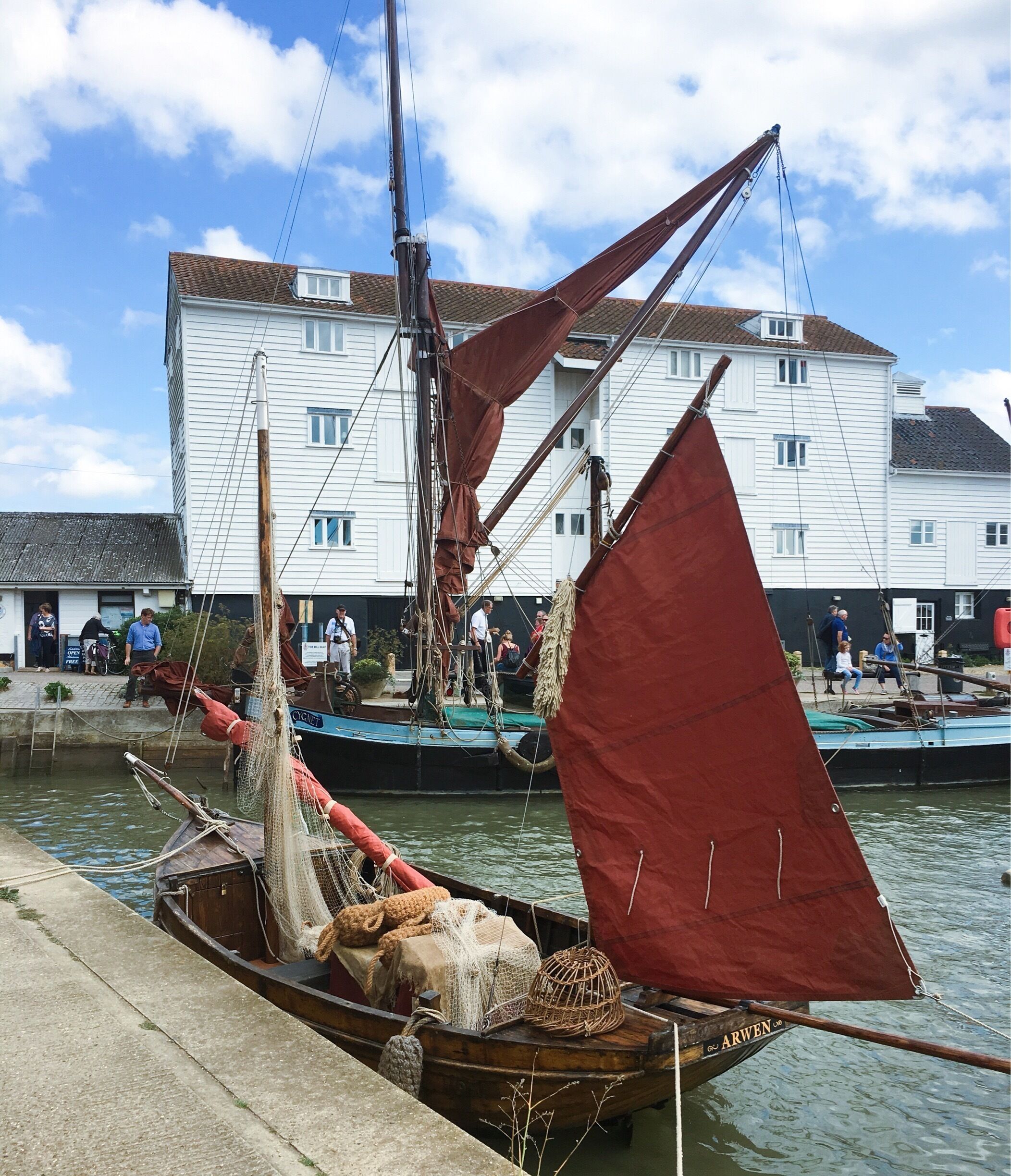 A traditional sailing boat docked at a pier with a red sail, surrounded by buildings and a clear sky.