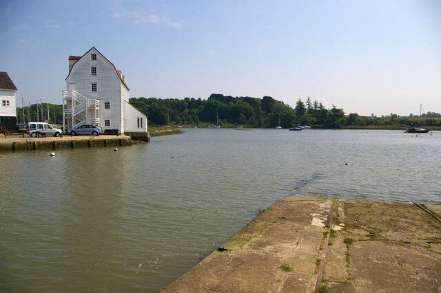 A calm river with a white building and parked cars on the left, surrounded by greenery and boats in the distance.