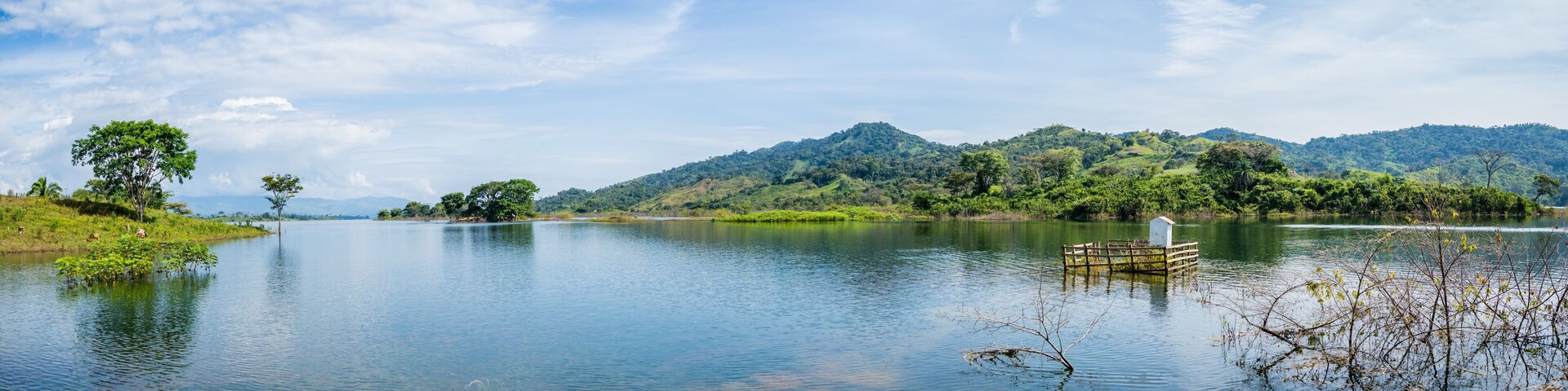 A calm lake with a wooden dock, surrounded by lush greenery and hills.