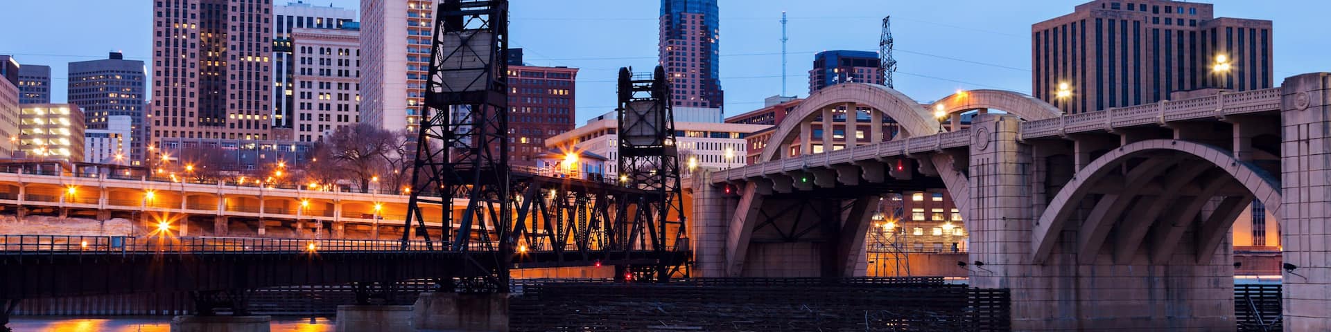 A city skyline at dusk with a prominent bridge reflecting city lights on the water.
