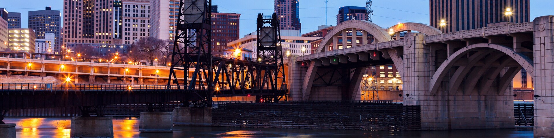Horizonte urbano al atardecer con un puente destacado que refleja las luces de la ciudad en el agua.