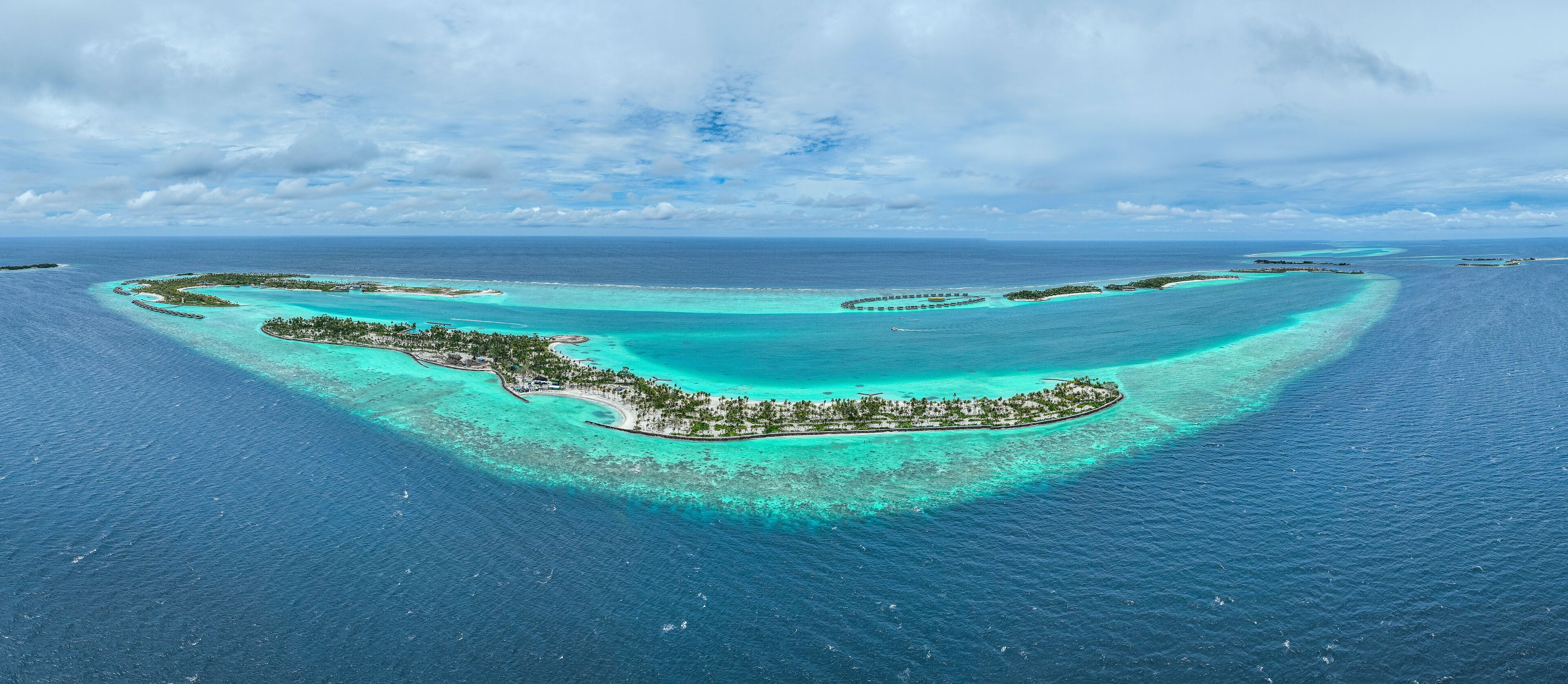 An aerial view of a tropical island with clear turquoise waters, surrounded by a coral reef, and a small settlement with palm trees.