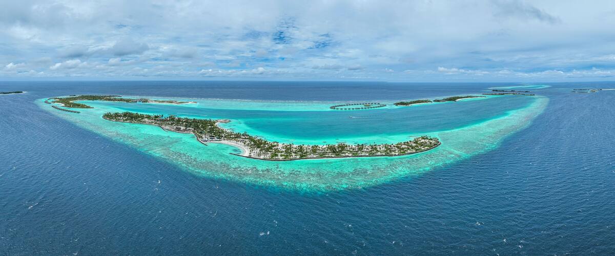 An aerial view of a tropical island with clear turquoise waters, surrounded by a coral reef, and a small settlement with palm trees.