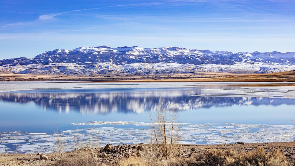 A snow-covered mountain range, a calm lake with ice, and sparse vegetation in the foreground.