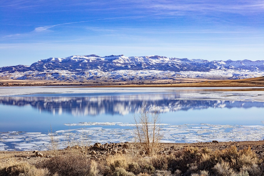 A snow-covered mountain range, a calm lake with ice, and sparse vegetation in the foreground.