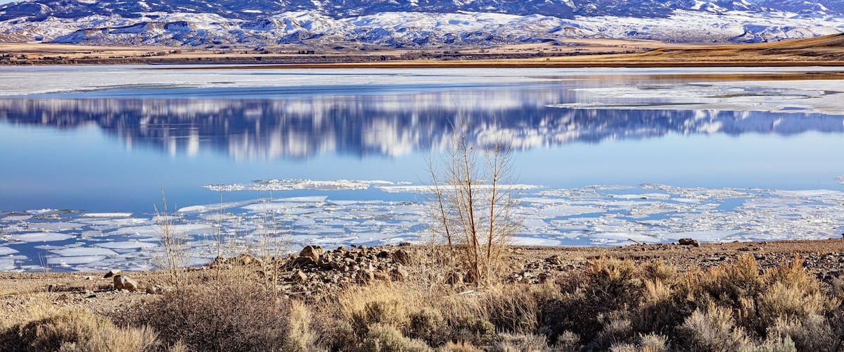 Buffalo Bill Cody Reservoir and snow-covered Carter Mountain Range during spring with ice in the water and blue cloudy sky during spring in Wyoming, USA.