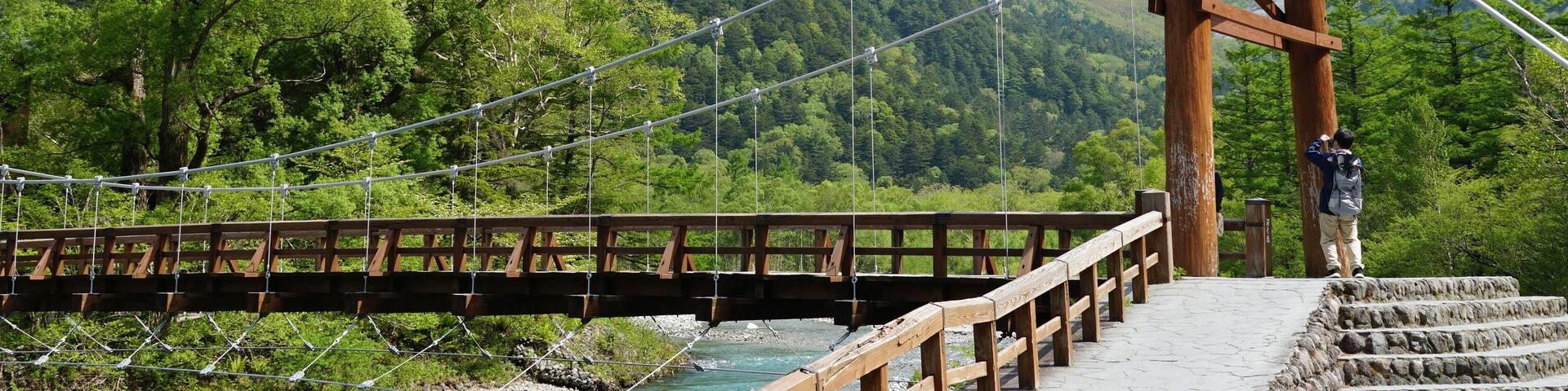 A suspension bridge with a wooden walkway, surrounded by lush green trees and snow-capped mountains in the background.