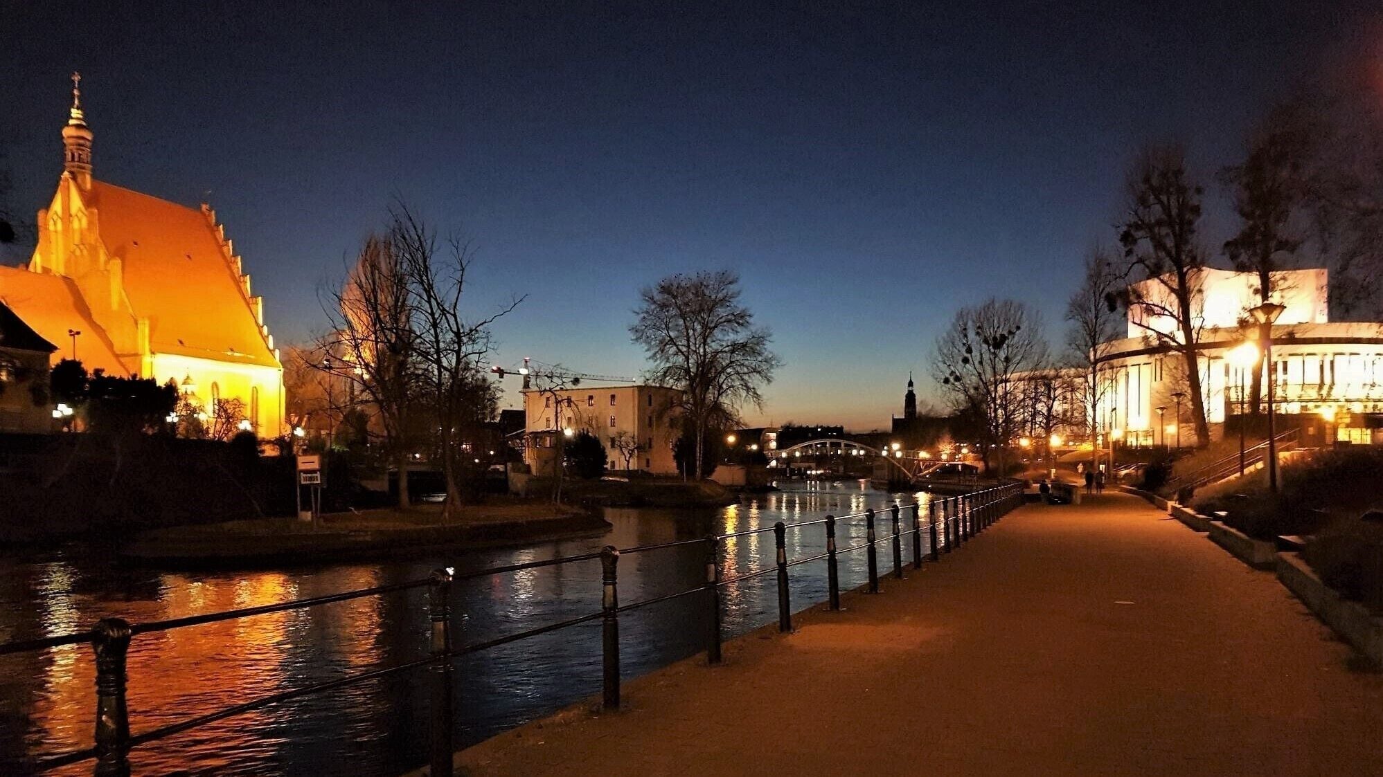 A riverside walkway at dusk with illuminated buildings and a church spire.