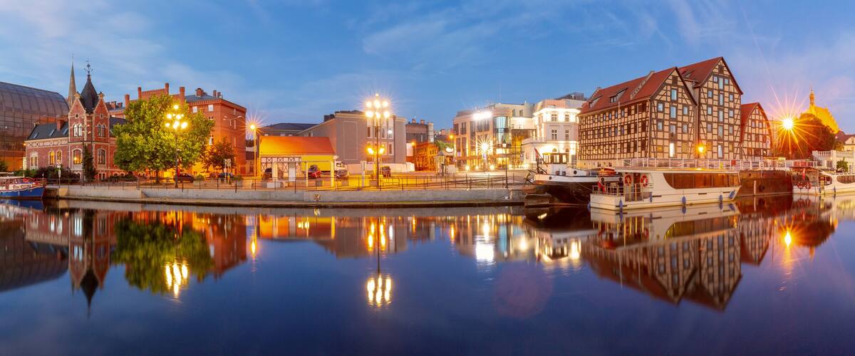 A riverside cityscape with historic buildings, a modern glass structure, and a traditional timber-framed house.