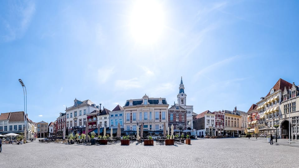 A cobblestone square with historic buildings, a prominent clock tower, and a large curved building with flags.