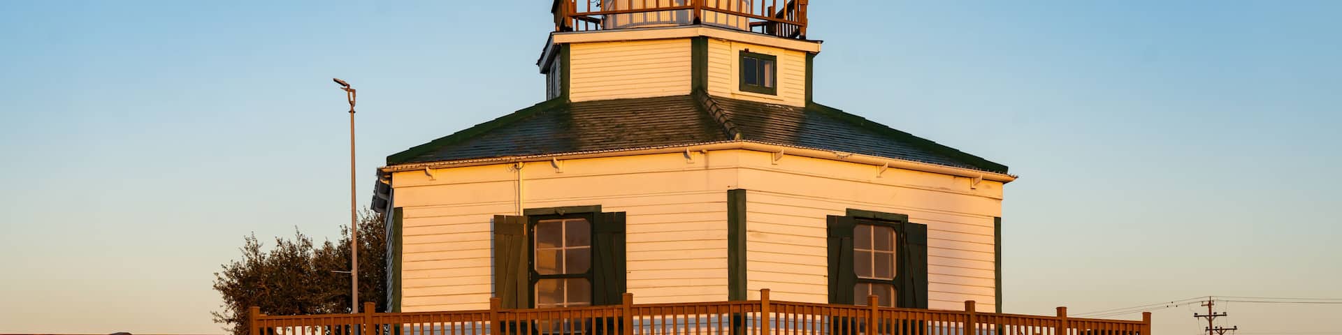 A lighthouse with a wooden deck and a green roof, surrounded by a wooden fence and flowering bushes.