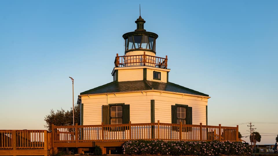 A lighthouse with a wooden deck and a green roof, surrounded by a wooden fence and flowering bushes.