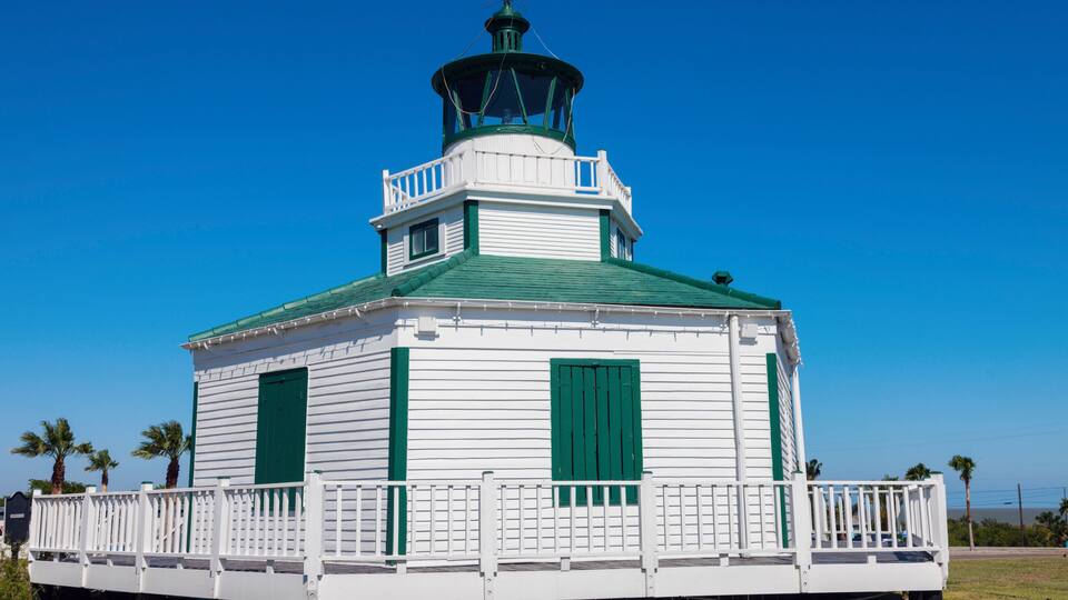 A white lighthouse with green trim and a green roof, surrounded by a white railing, under a clear blue sky.
