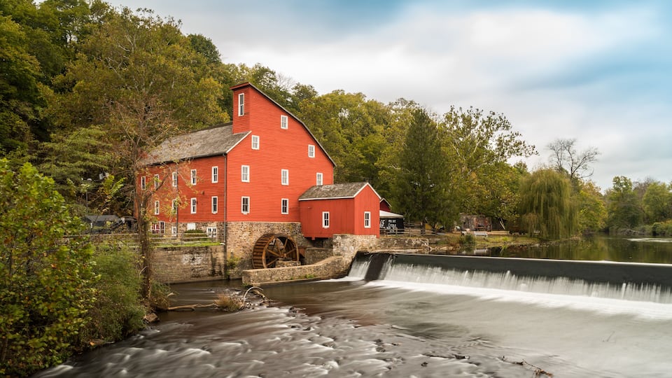A red mill with a water wheel by a river, surrounded by trees and greenery.