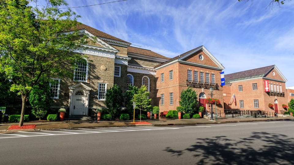 A historic brick building with arched windows and a central entrance, surrounded by trees and a clear blue sky.