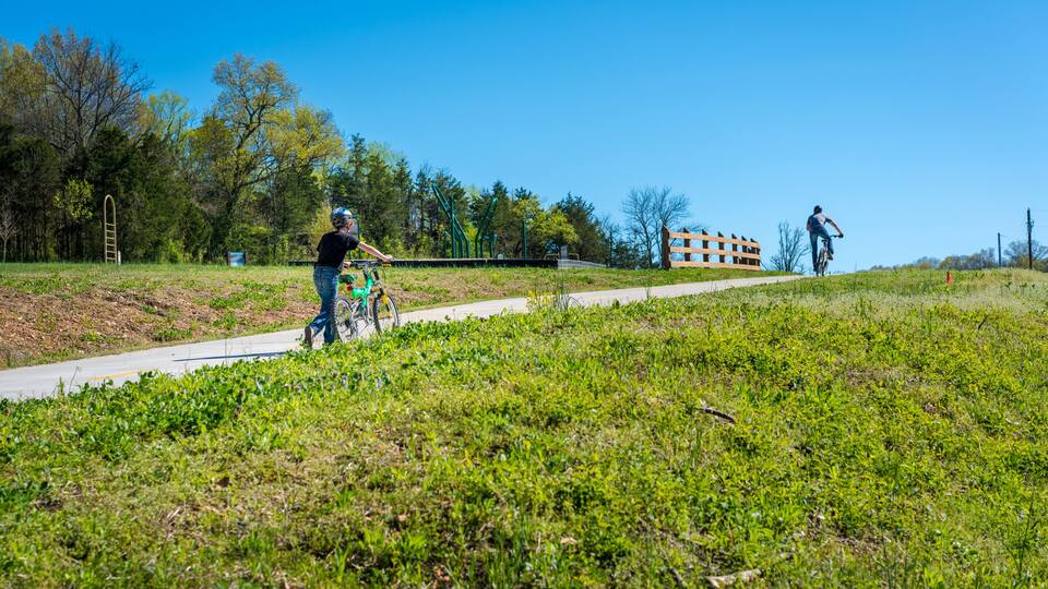 A park with a paved path, green grass, trees, and a distant building.