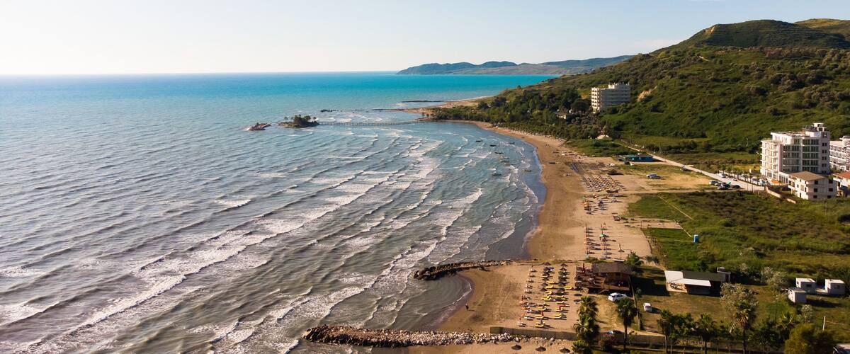 A coastal beach with waves, a sandy shore, and a hilly landscape with buildings.