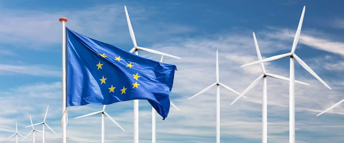 A European Union flag waving in front of wind turbines under a blue sky.