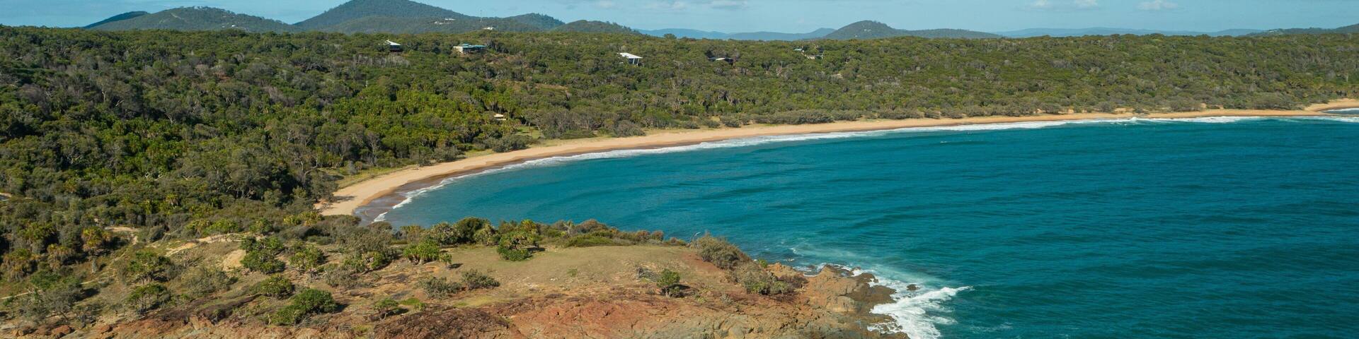 A coastal landscape with a beach, rocky outcrops, and a forested hill.