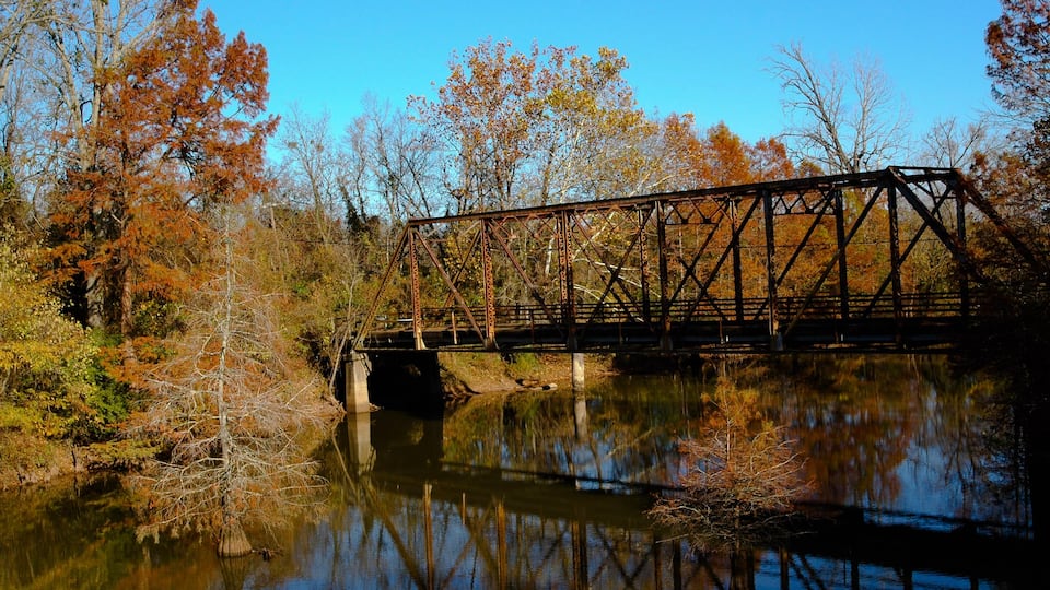 A rusted metal bridge over a calm river, surrounded by autumn-colored trees.