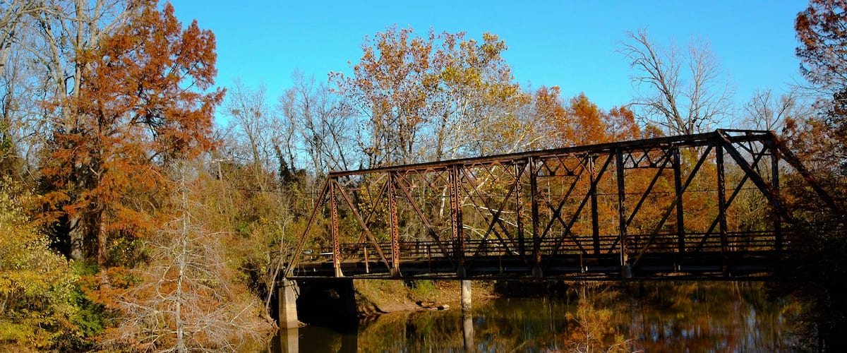 A rusted metal bridge over a calm river, surrounded by autumn-colored trees.