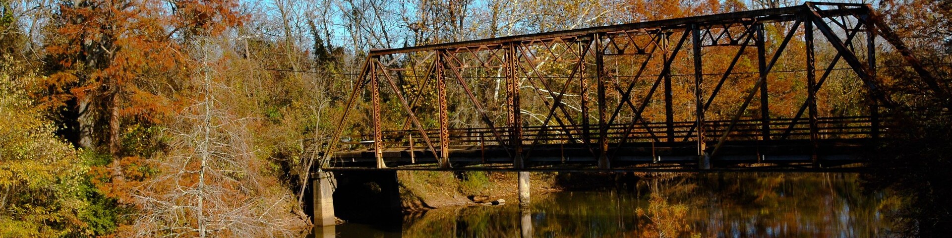 A rusted metal bridge over a calm river, surrounded by autumn-colored trees.