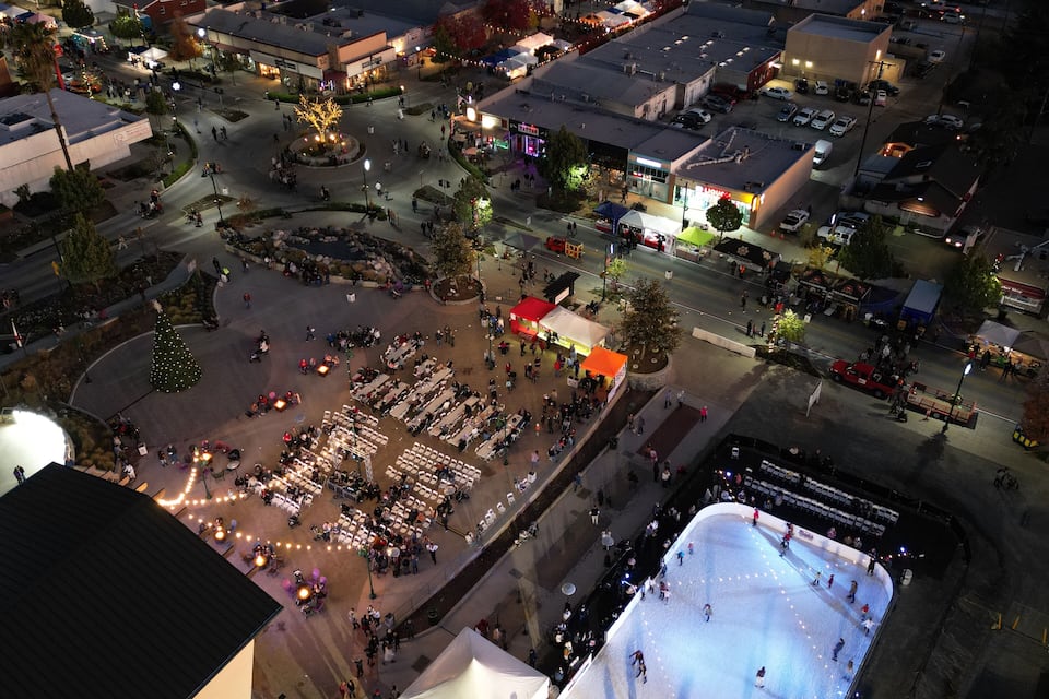 An aerial view of a city square at night with a large ice rink, outdoor seating areas, and surrounding buildings.
