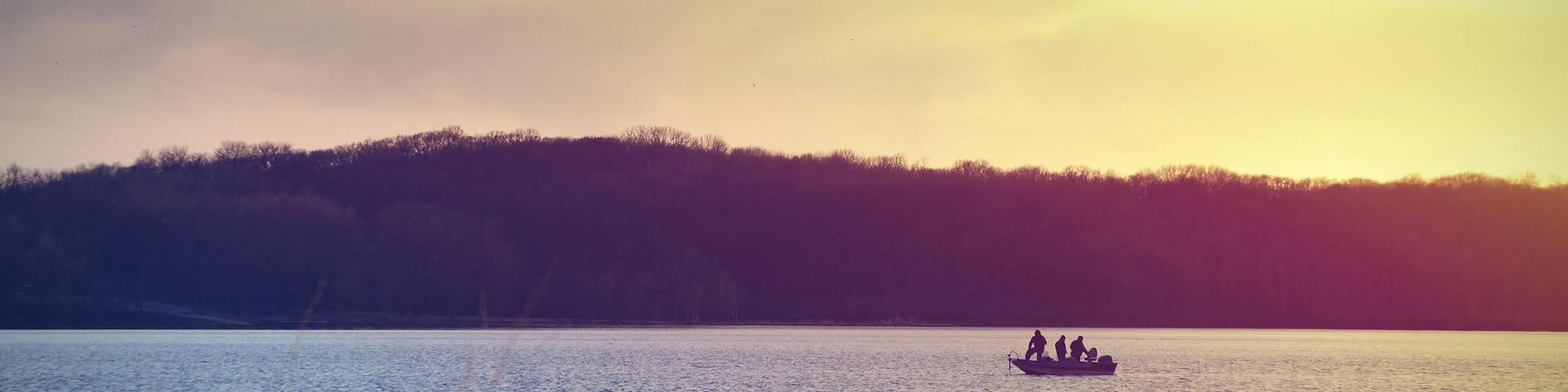 A calm lake with a boat in the distance and a forested hill in the background.