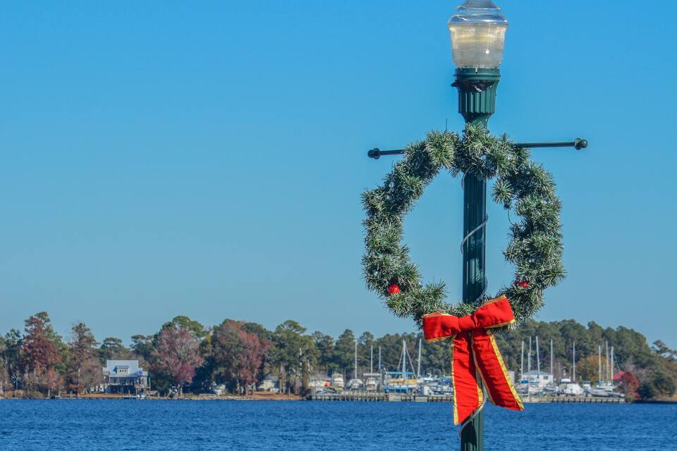 Un muelle con un farol decorativo, una barandilla, y un fondo de árboles y barcos.