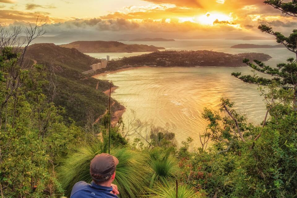 Un homme est assis sur un rocher, d’où il domine un paysage côtier composé de collines et d’une étendue d’eau, au moment du coucher du soleil.