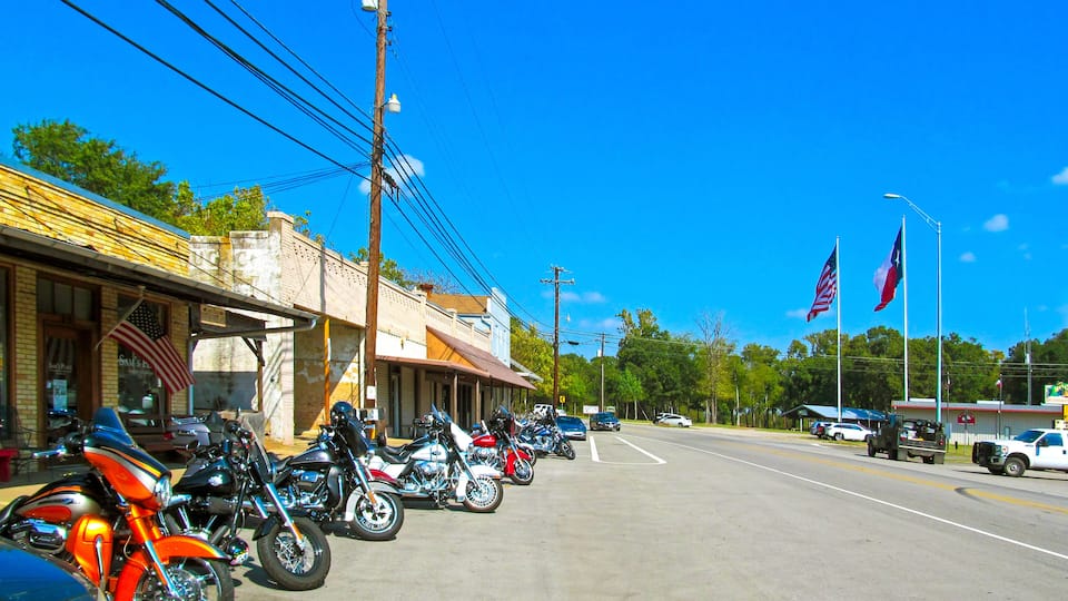 A street lined with parked motorcycles and cars, a building with an American flag, and a Texan flag.