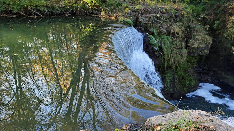 A waterfall cascading over a rocky ledge into a calm river surrounded by lush greenery.