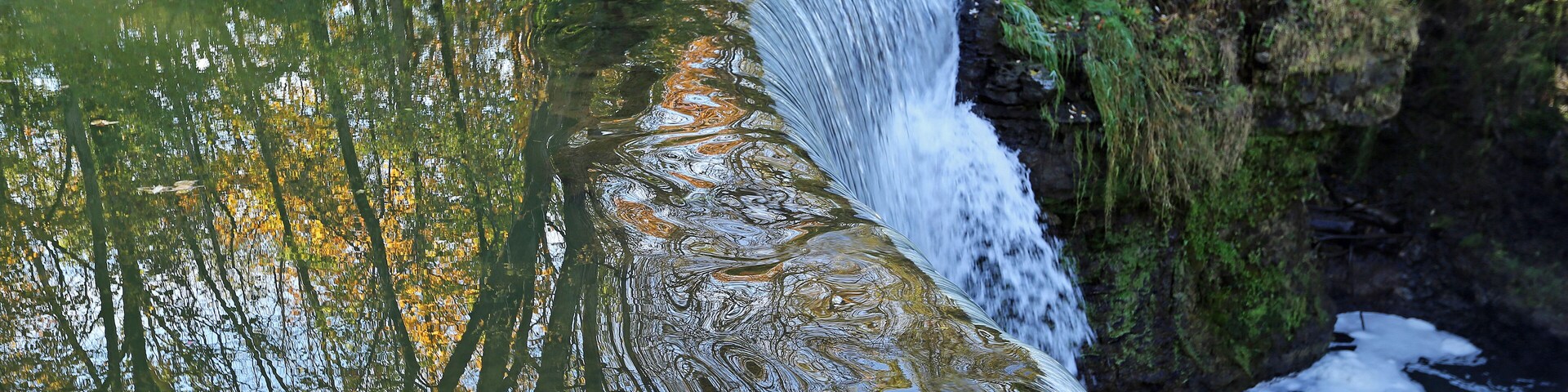 Una cascada se precipita sobre un saliente rocoso hacia un río tranquilo rodeado de exuberante vegetación.