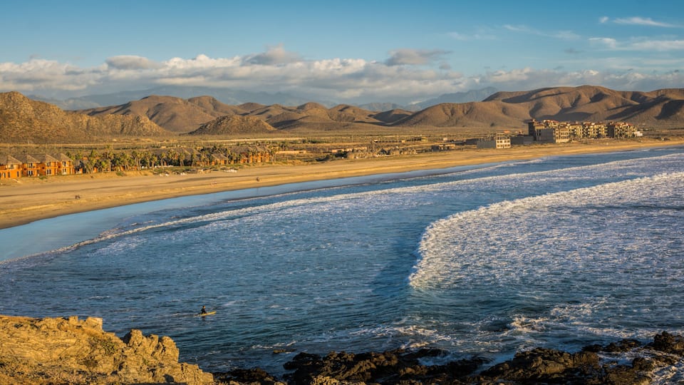 A coastal landscape with a beach, rocky shoreline, and distant mountains.