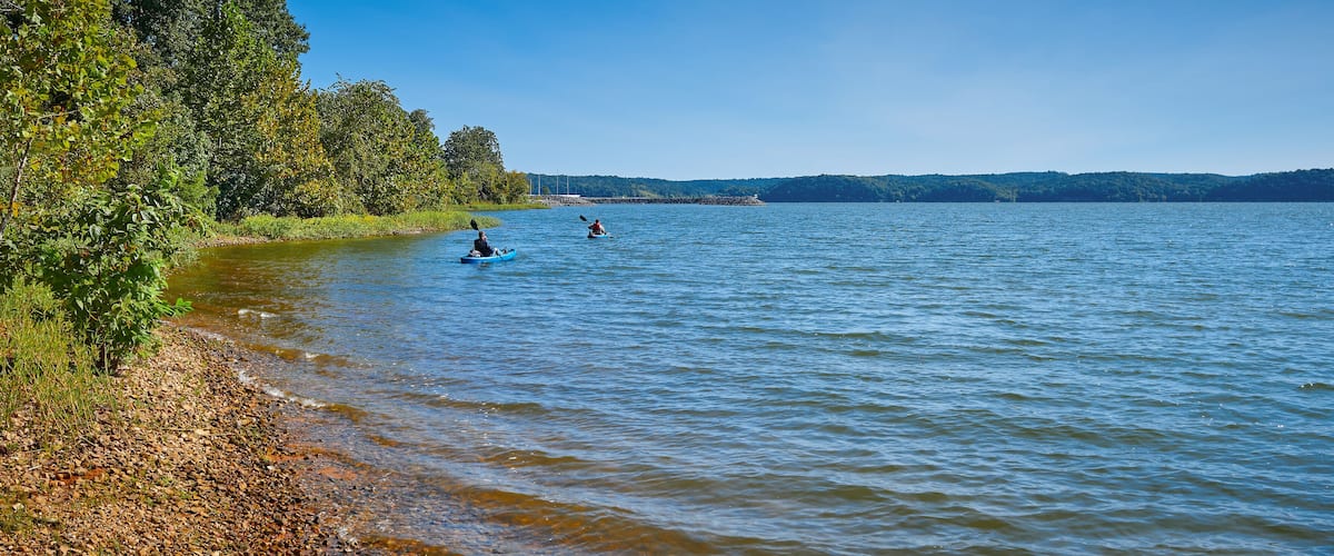 A calm lake with kayakers, surrounded by dense trees and a distant shoreline.