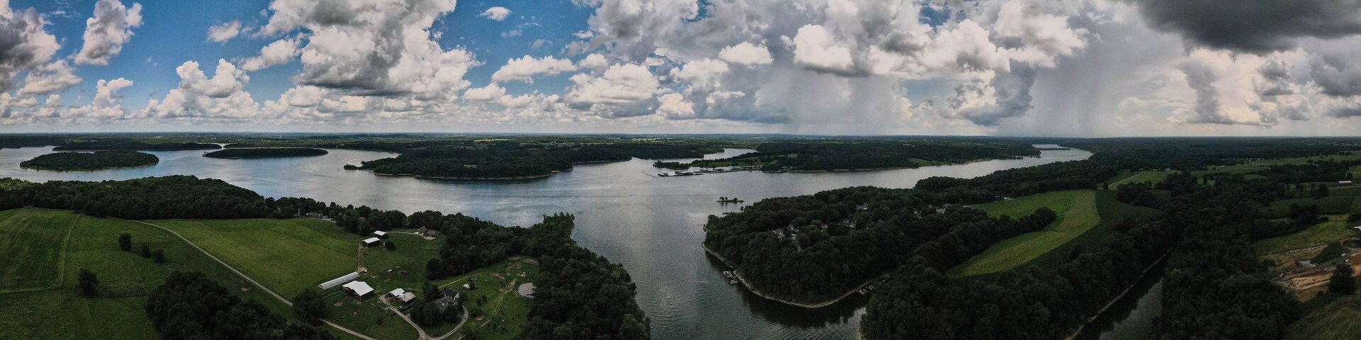 Un río rodeado de campos verdes y pequeños edificios, con grandes nubes en el cielo.
