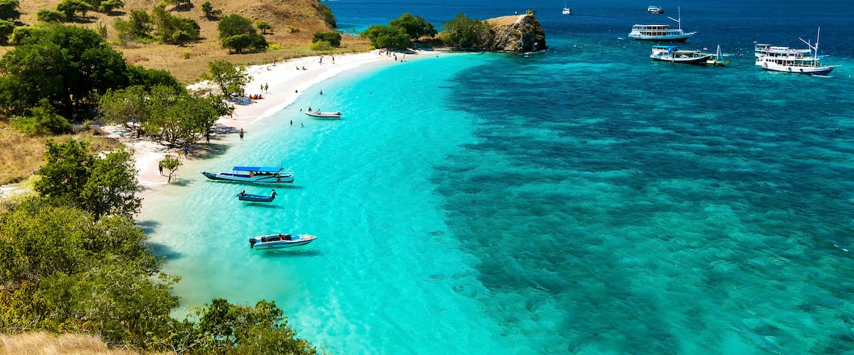 A coastal landscape with clear turquoise waters, sandy beach, boats, and hilly terrain.