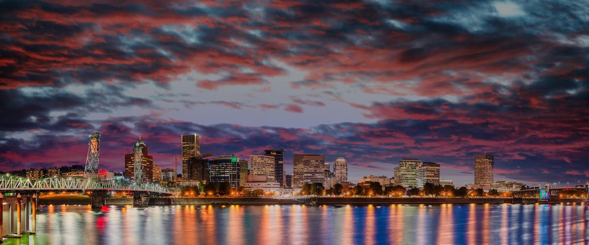 A city skyline with a river in the foreground, a bridge, and a colorful sky.