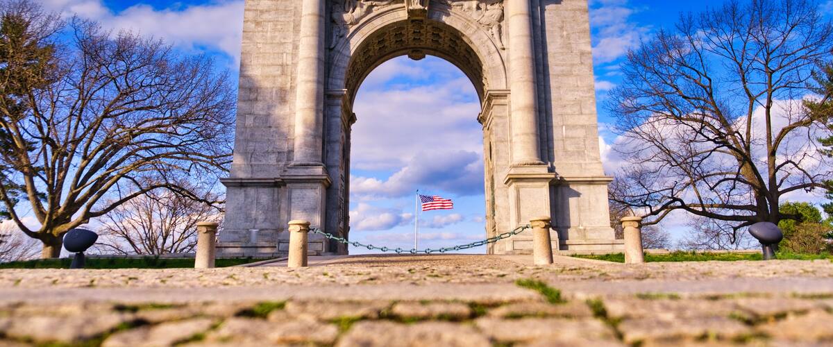 A stone archway with an American flag, surrounded by bare trees and a clear sky.