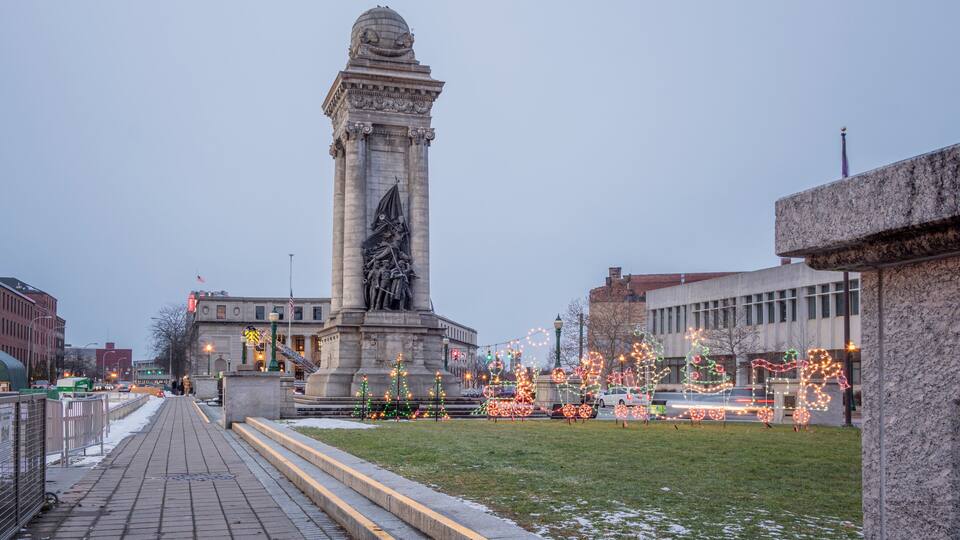 A monument with a statue, surrounded by a paved walkway and decorated with holiday lights.